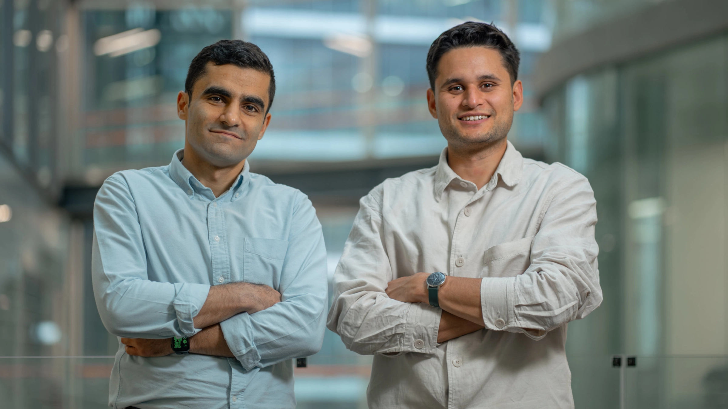 Two men standing side by side, one in light blue button-up, one in beige, in modern office setting.