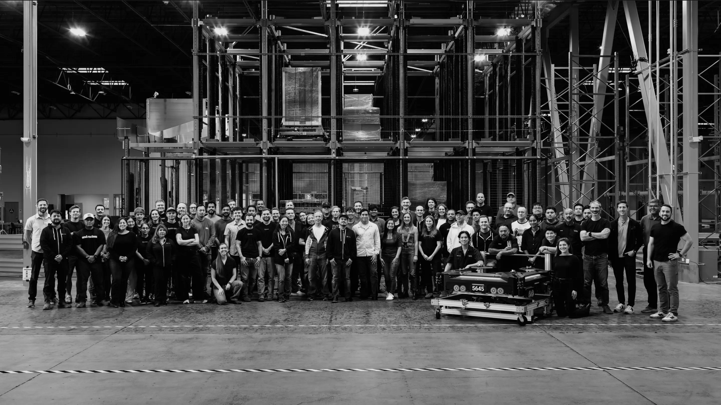 Large group of people standing together in an industrial warehouse setting with metal framework visible above them.