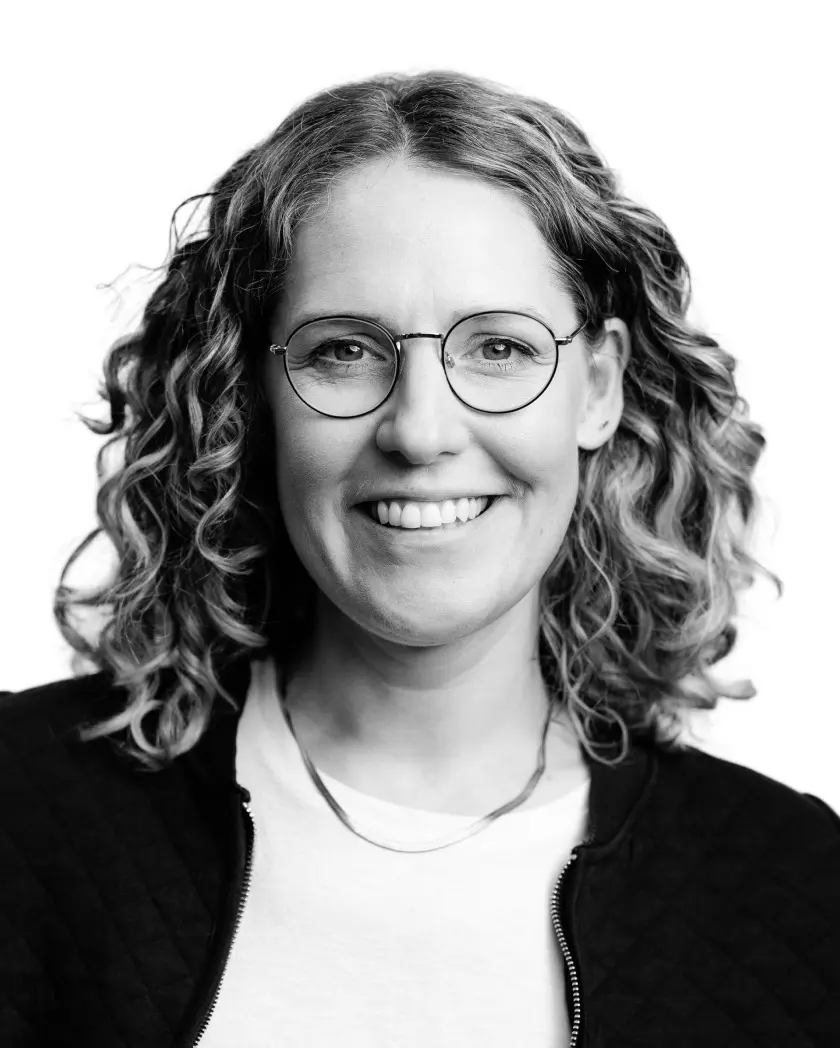 Black and white headshot portrait of a woman with curly shoulder-length hair and round glasses, smiling.