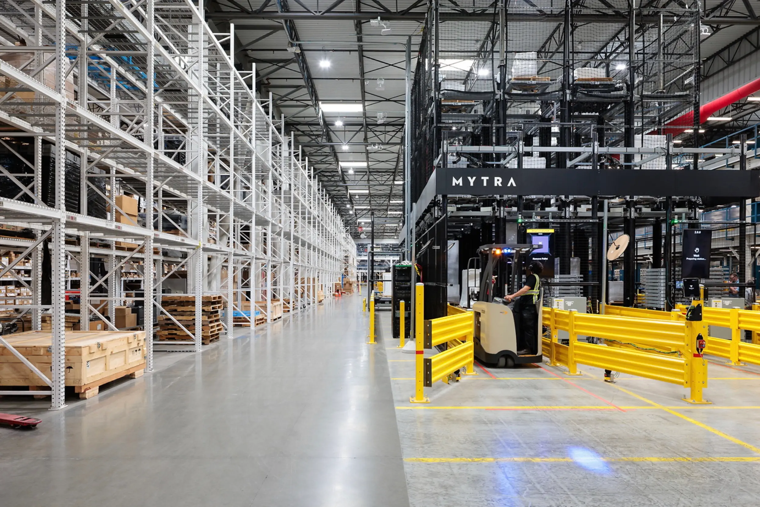 Modern warehouse interior with yellow safety barriers, shipping containers, and people working in logistics facility.